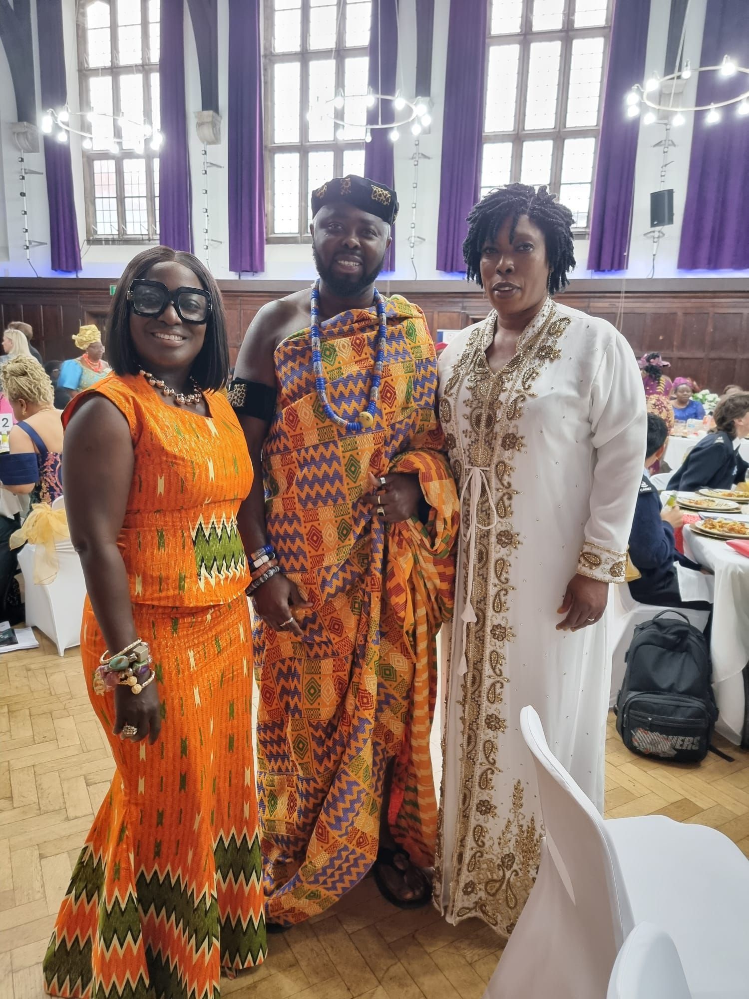 Three people in traditional African attire pose indoors. A man in orange and blue fabric stands between two women, one in an orange dress and the other in white.
