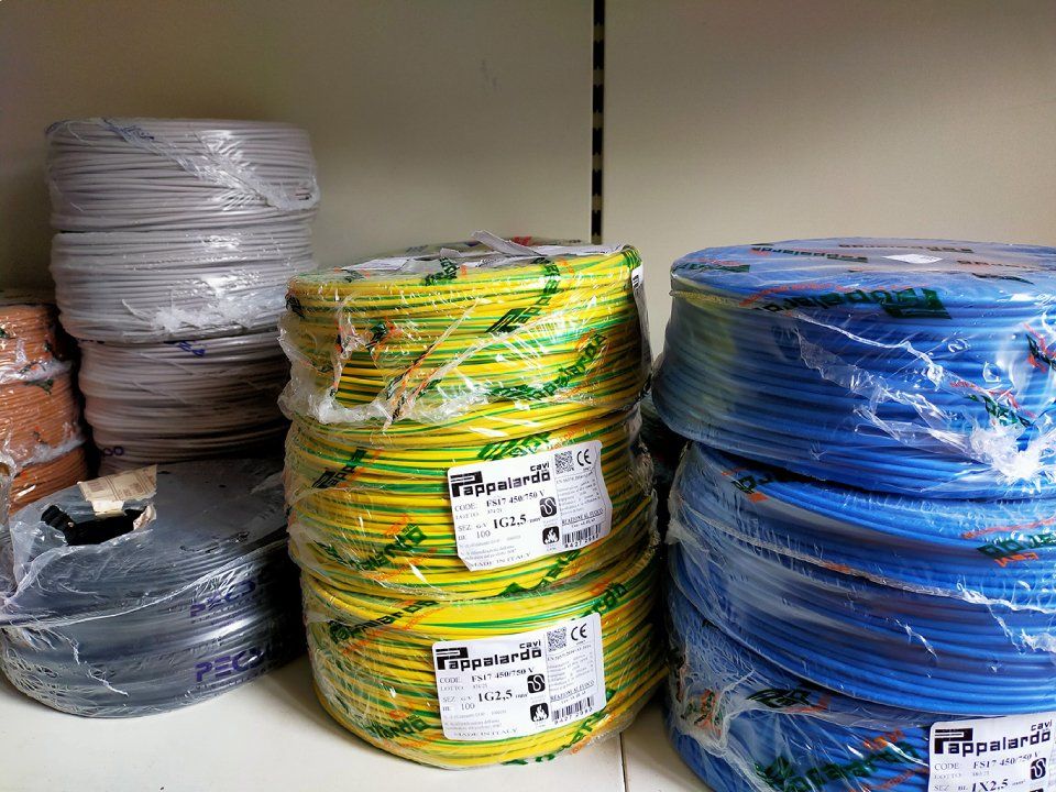 Pile di cavi arrotolati di colore grigio, giallo e blu, avvolti nella plastica, sugli scaffali di un magazzino.