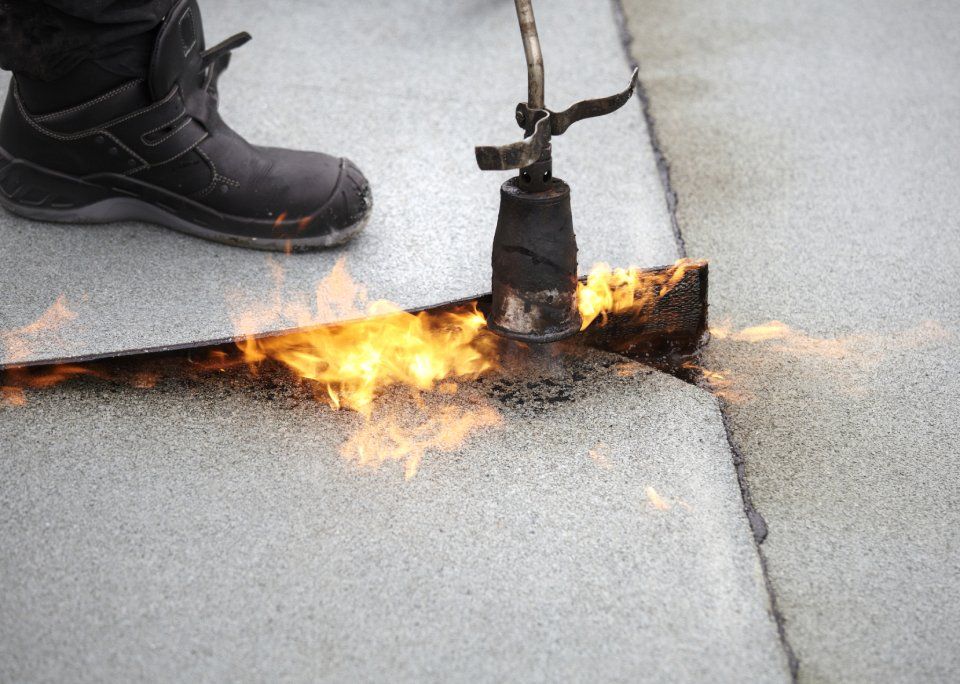 Una fiamma ossidrica fonde una giuntura in una membrana grigia del tetto accanto allo stivale nero di un operaio.