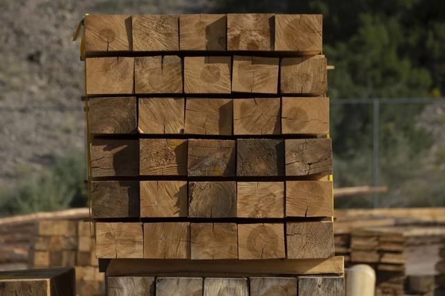 Travi di legno accatastate in un cantiere edile all'aperto, con altre cataste di legno sfocate sullo sfondo.