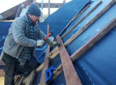 Worker nailing wooden planks onto a blue tarp-covered roof frame