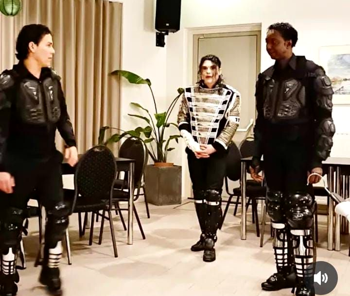Drie artiesten staan ​​op het podium en dansen in zwarte outfits met zilveren accenten, onder roze en rode podiumverlichting.