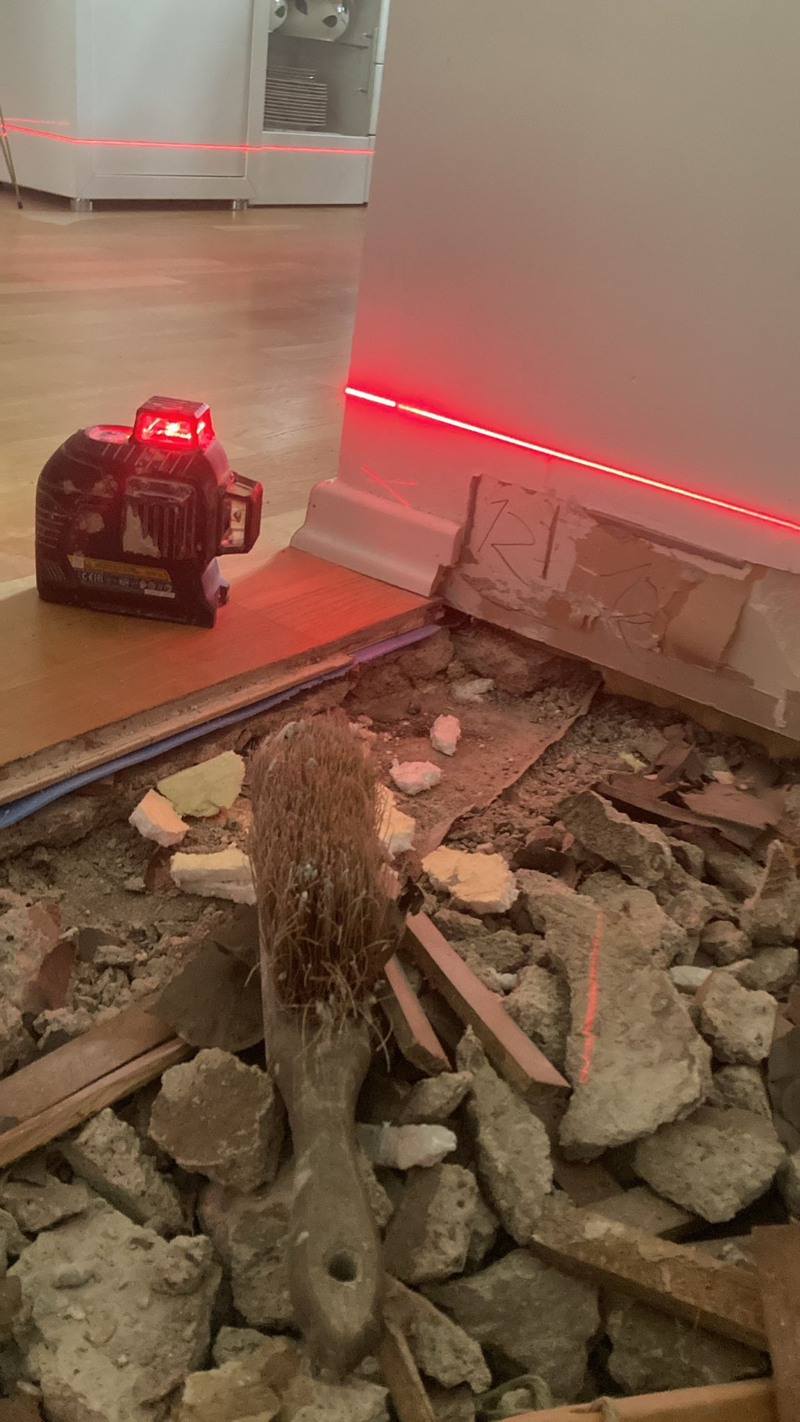 Laser level projects a red line on a wall during floor demolition. Debris and a brush are in the foreground.