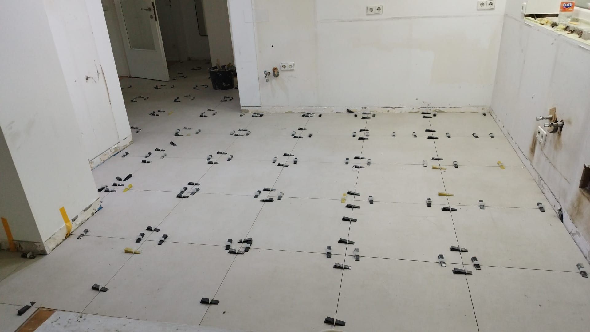 Floor tile installation in progress; white tiles laid out with leveling clips and spacers, in a room with white walls.