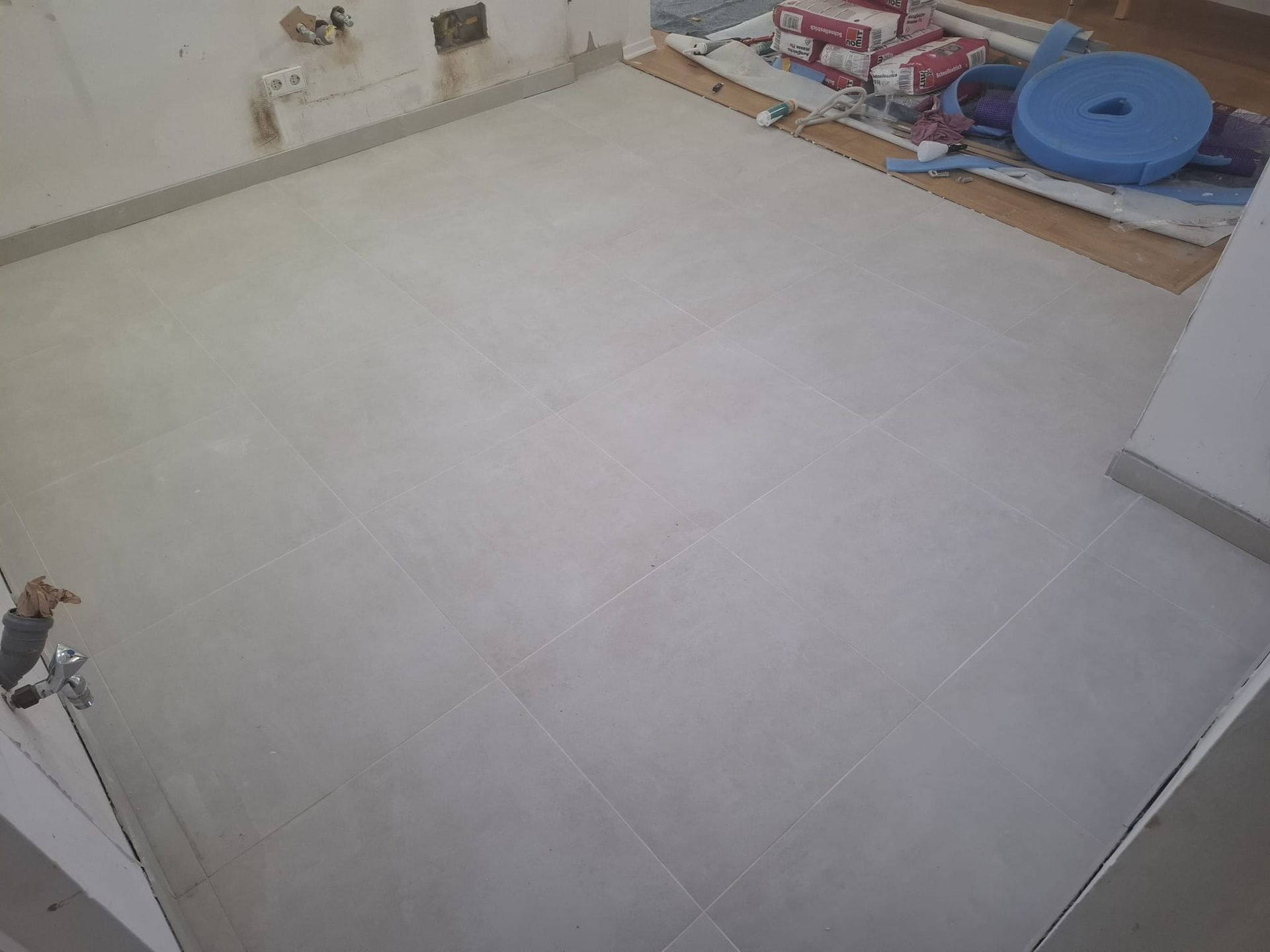 Light gray tiled floor in a room, partially installed, with construction materials visible in the background.
