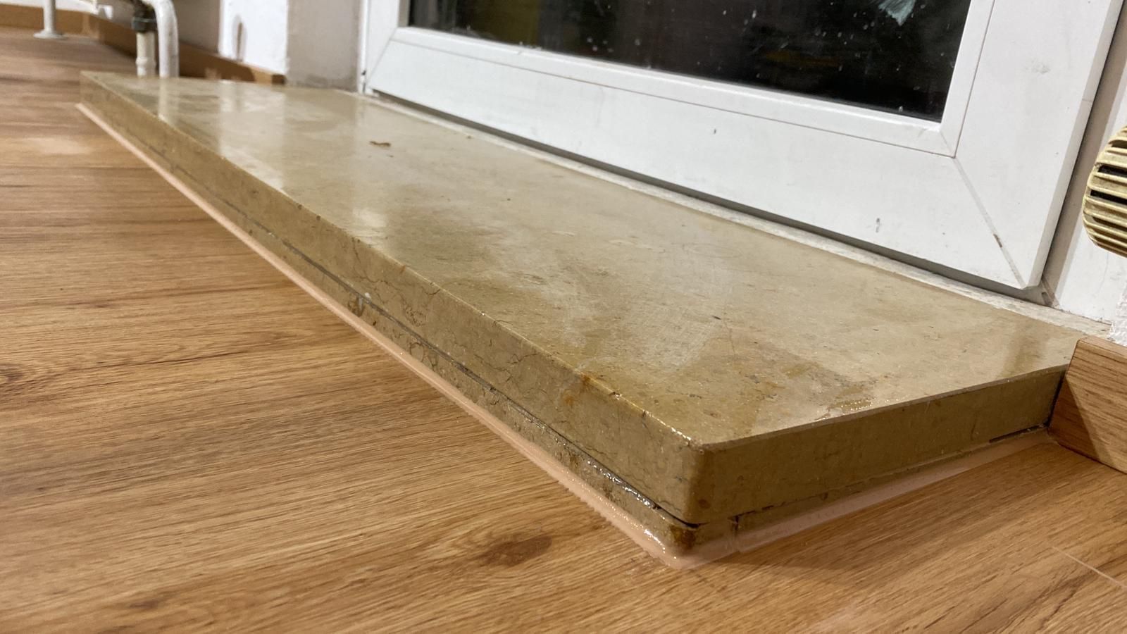 A window sill made of light-colored material over a wooden floor, next to a white window frame.