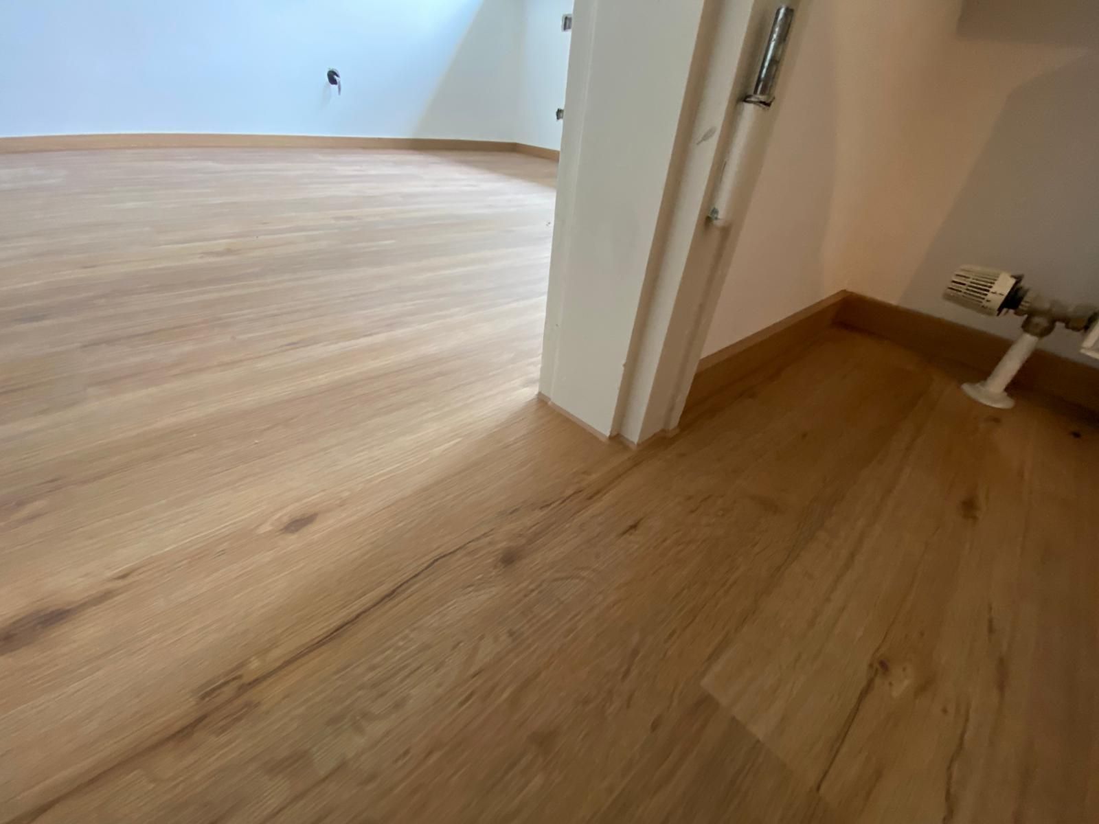 Wooden flooring in a room with a doorway and a radiator. Light brown planks, neutral walls.