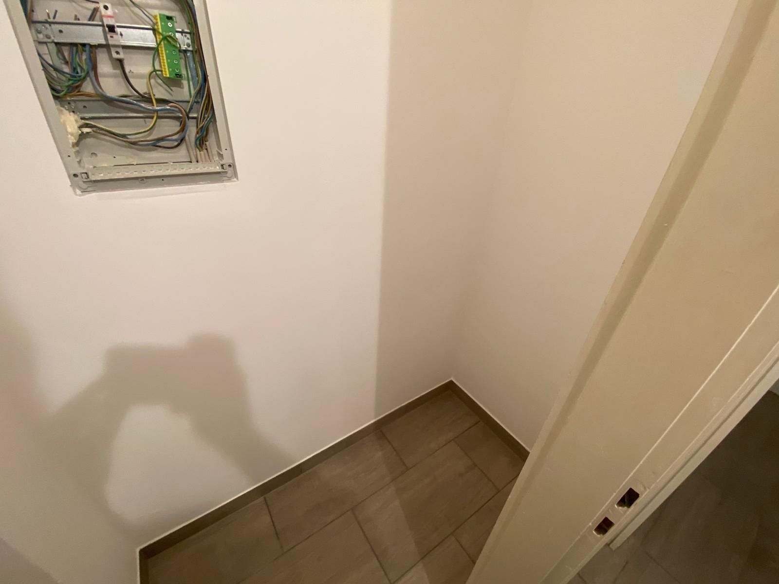 Small, empty room with beige tile floor, white walls, and an open electrical panel.
