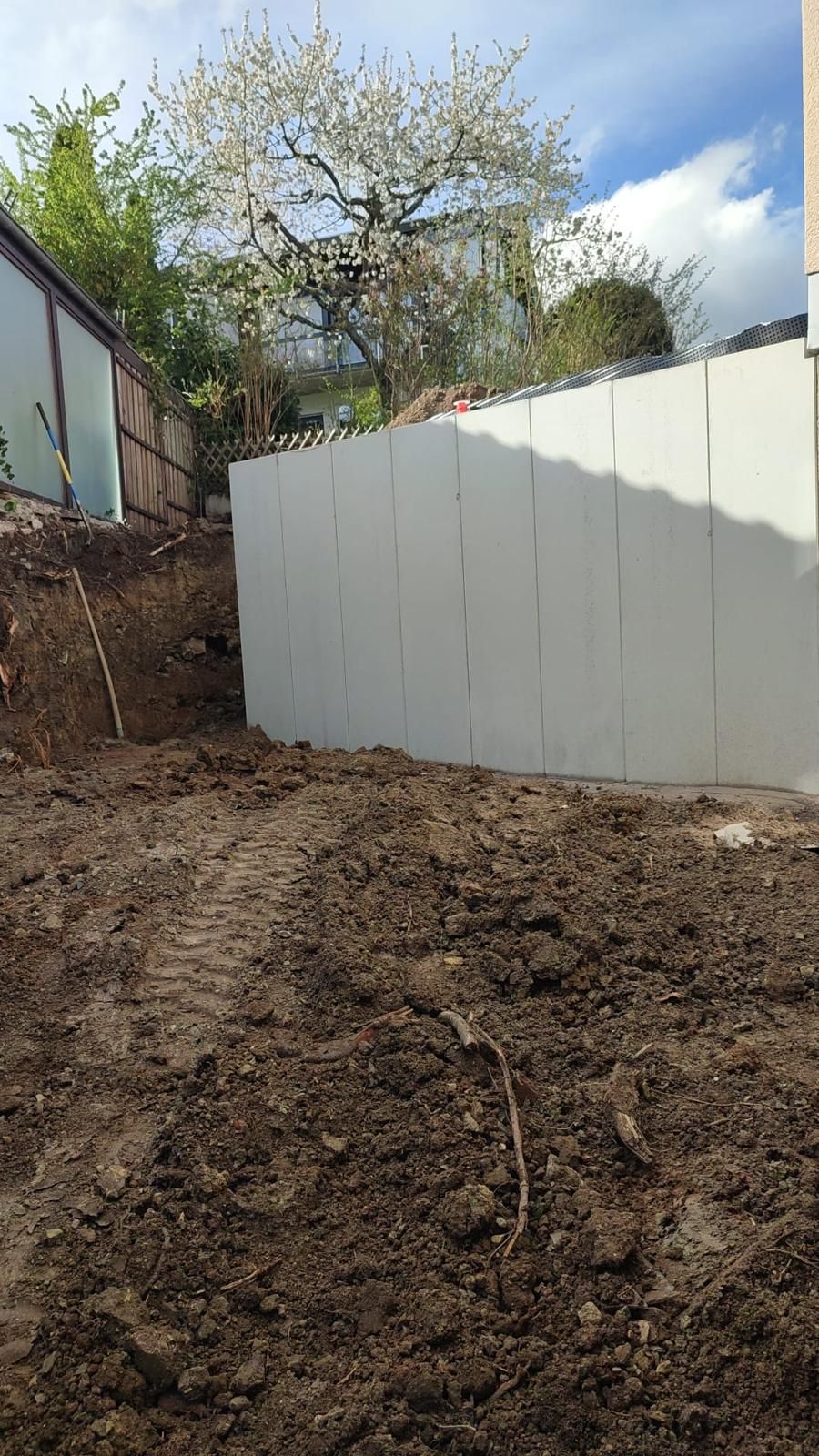 A newly built white retaining wall beside a muddy dirt embankment, under a blossoming tree.