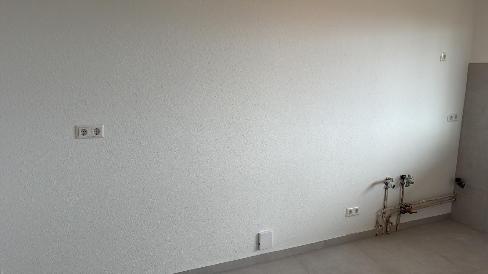 White textured wall with electrical outlets, water pipes, and a gray floor.