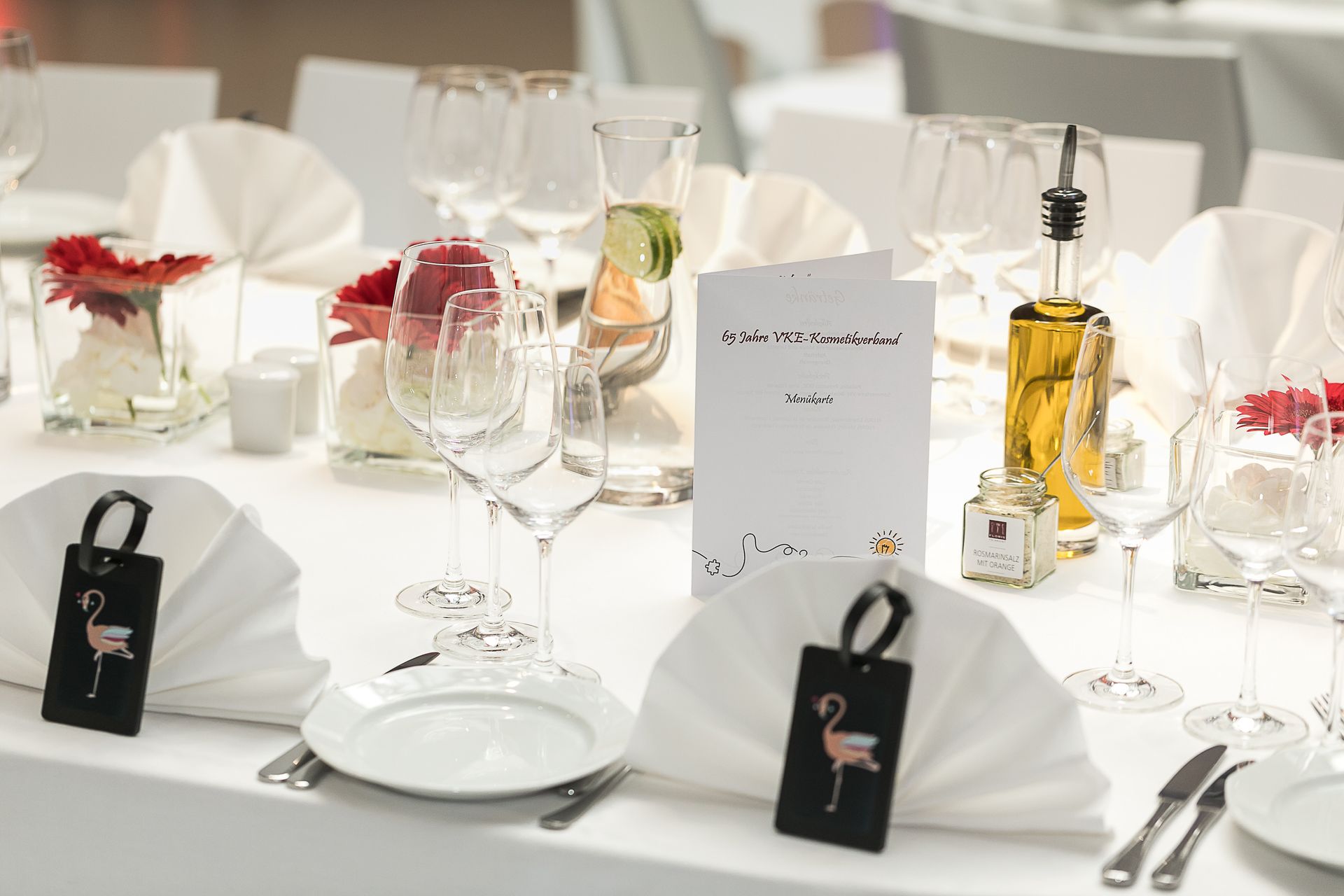 Eine formelle Hochzeitstafel mit weißer Tischwäsche, Tischkarten mit Flamingomotiven, Gläsern, einer Menükarte und einer Ölflasche.
