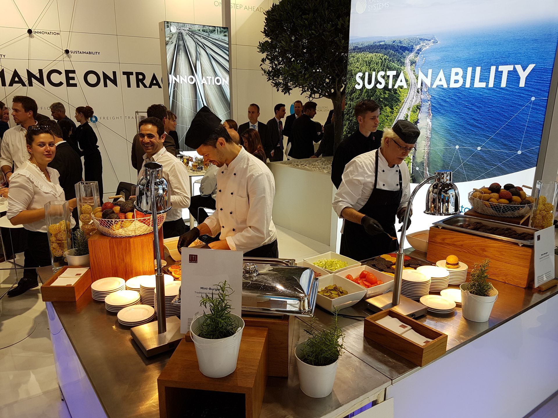 Professionelle Köche in weißen Uniformen bereiten Speisen an einer Cateringstation in einem hellen, modernen Veranstaltungsraum zu.