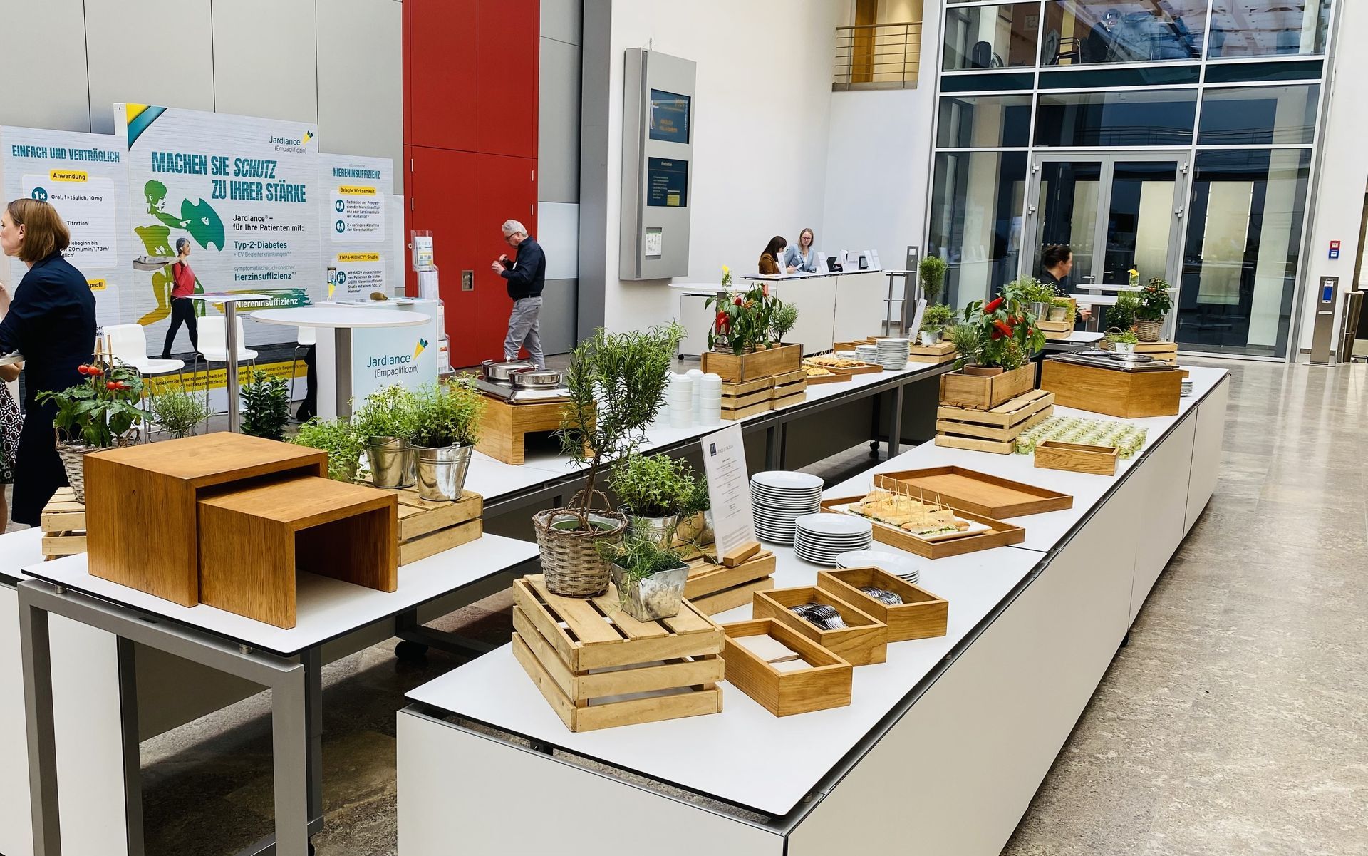 Catering-Auslage mit kleinen Topfpflanzen und hölzernen Servierkisten auf Tischen in einer hellen, modernen Atriumlobby.