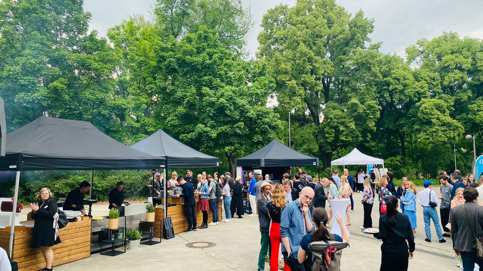 Bei einer Freiluftveranstaltung mischen sich die Menschen zwischen mehreren schwarz-weißen Pop-up-Zelten auf einem Kiesweg in der Nähe dichter grüner Bäume.