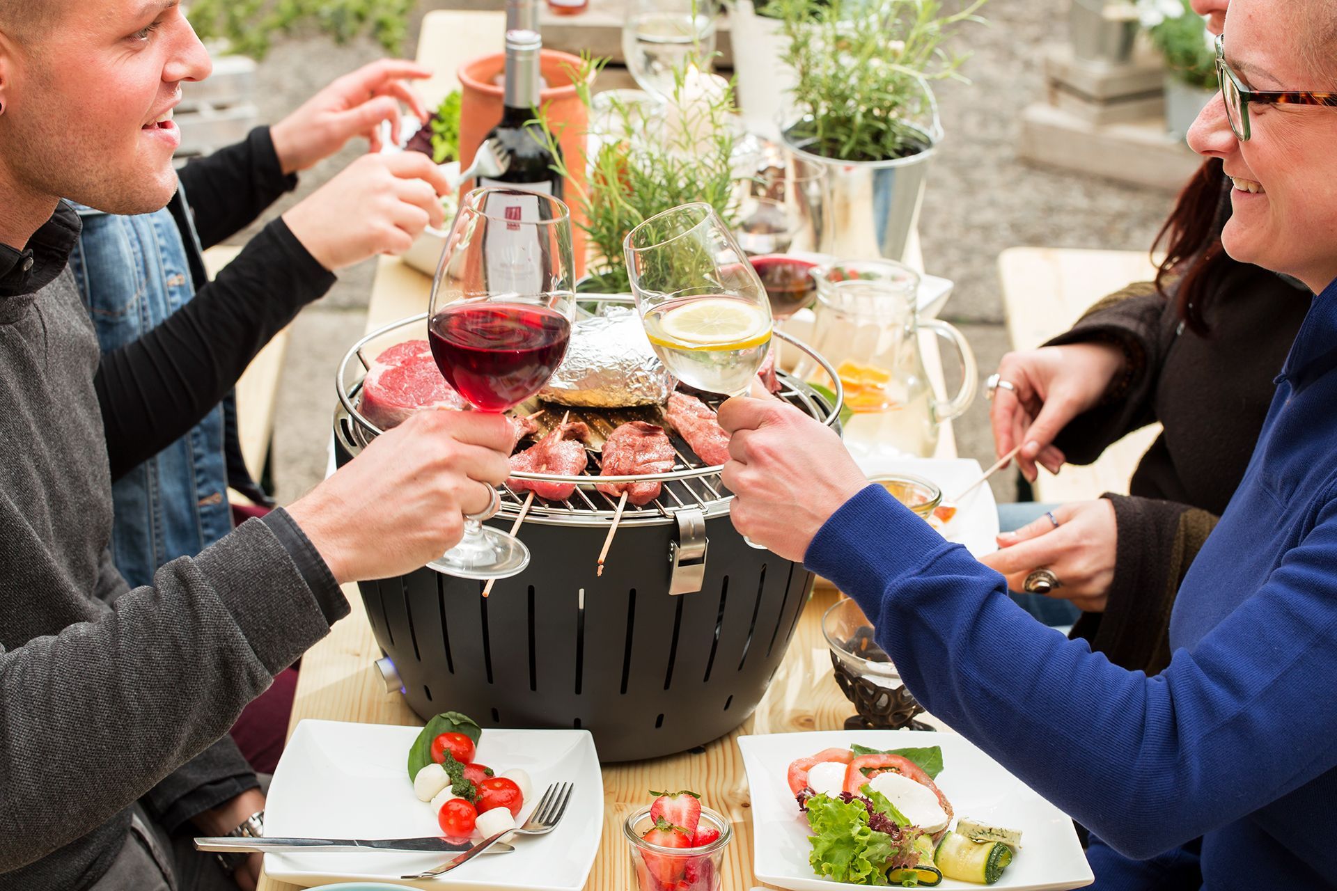 Menschen stoßen mit Wein und Getränken an, die über einem Tischgrill im Freien mit Tellern voller Salat und Brot serviert werden.
