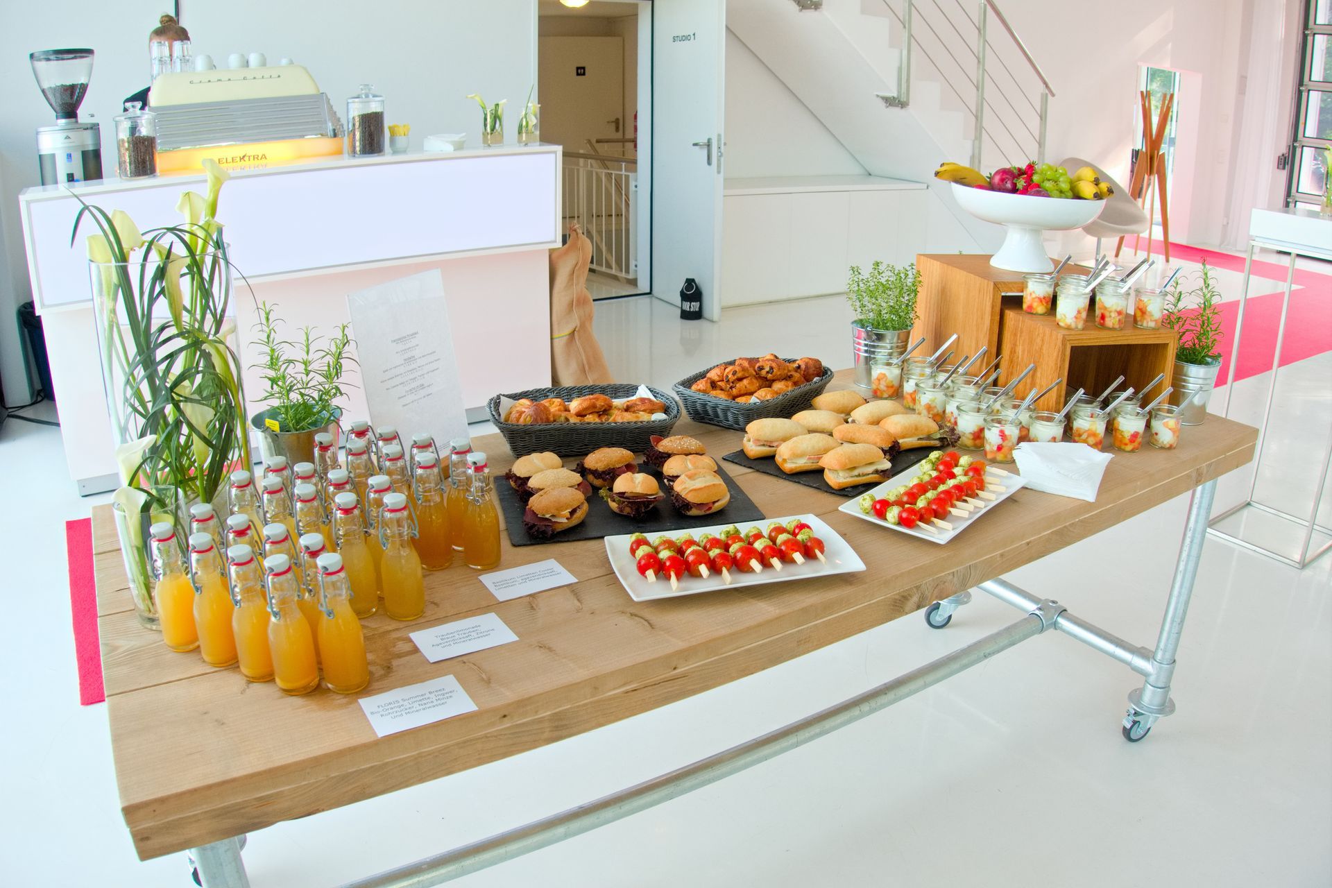 Ein Buffet mit Obst, kleinen Sandwiches, Vorspeisen und Reihen von abgefüllten Säften in einem hellen, modernen Veranstaltungsraum.
