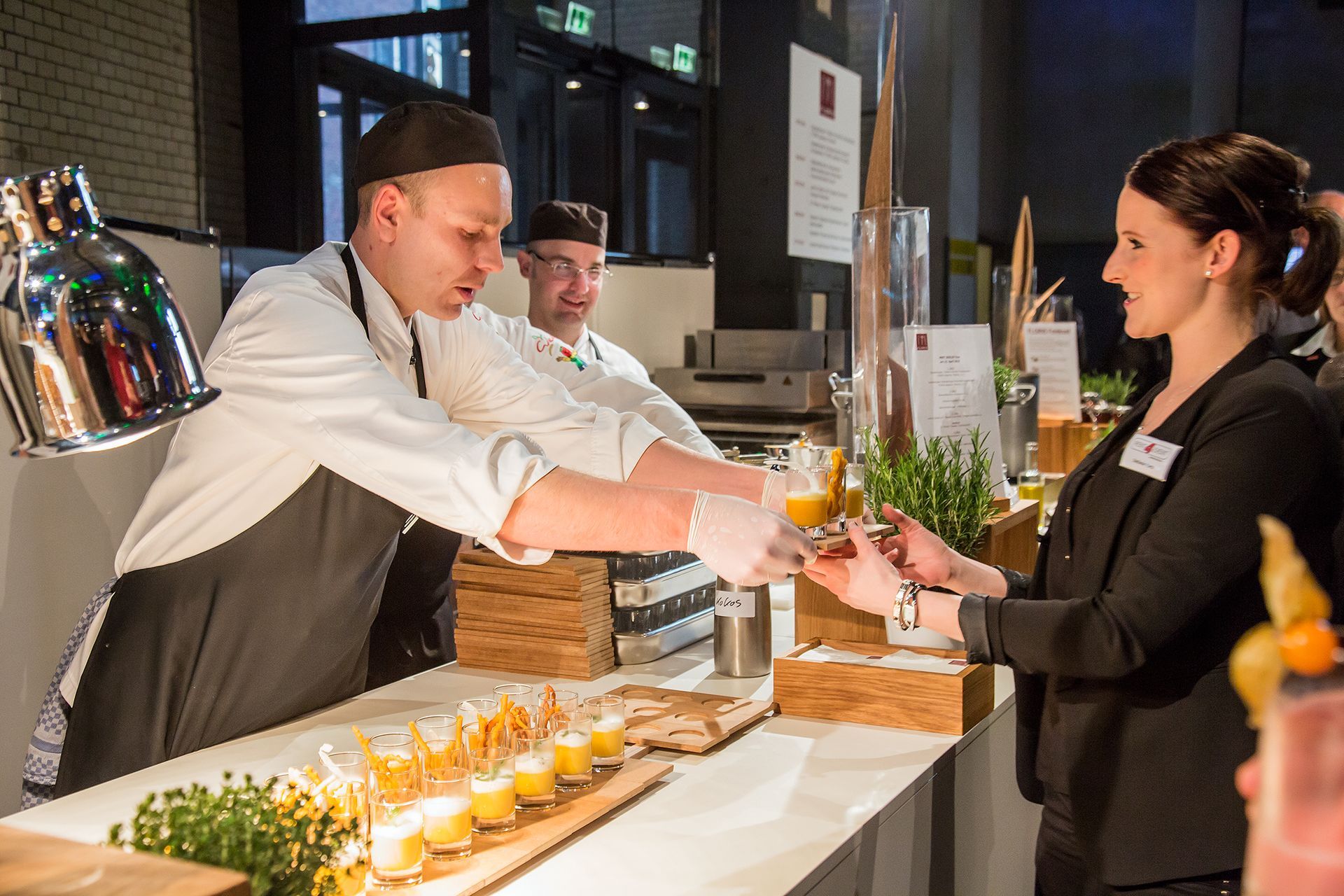 Bei einer Catering-Veranstaltung überreicht ein Koch in weißer Uniform einem Fachmann in schwarzem Blazer eine kleine Kostprobe.