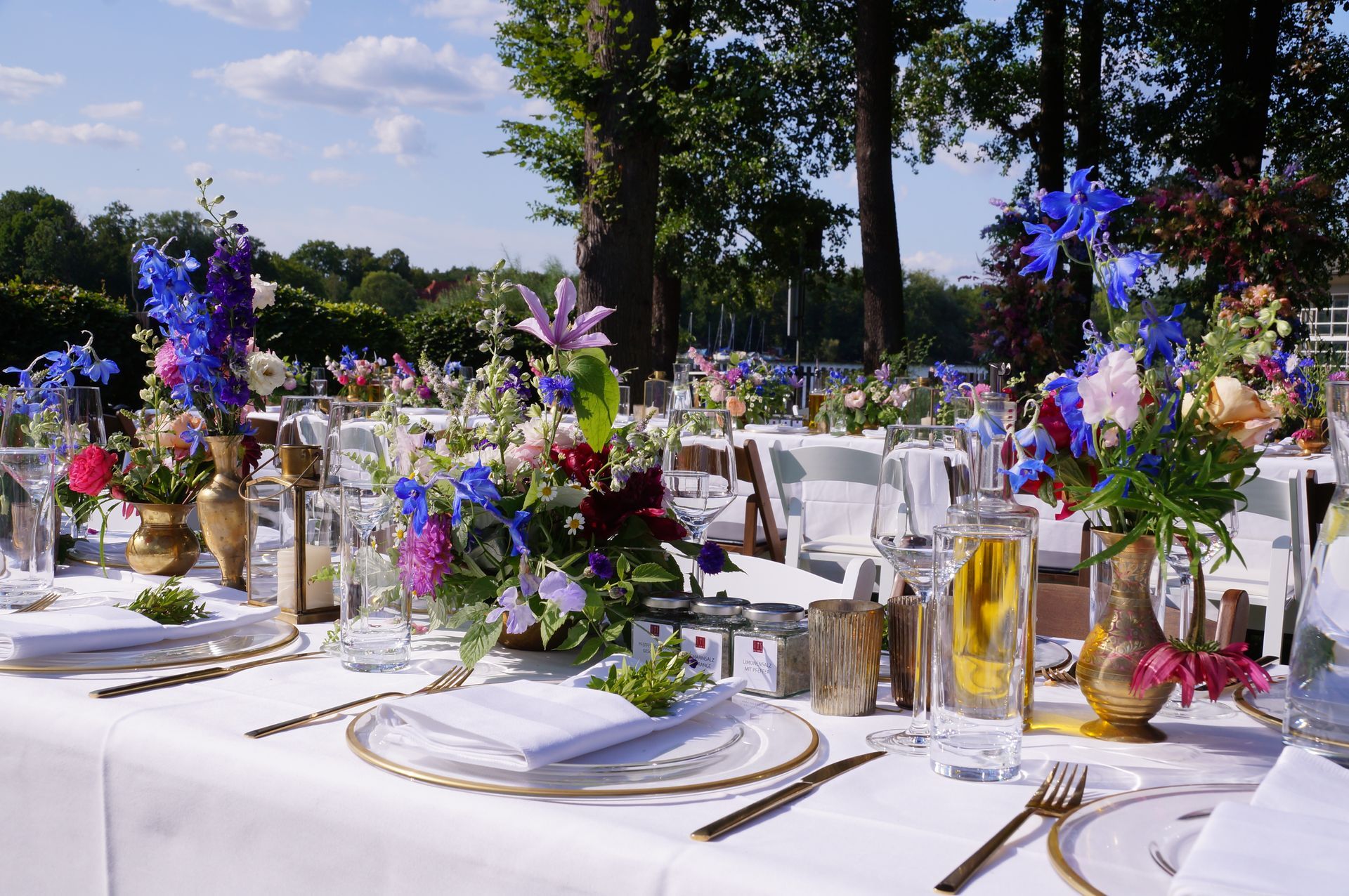 Ein langer, weiß gedeckter Esstisch für eine Veranstaltung im Freien, verziert mit leuchtend blauen und violetten Blumenarrangements.