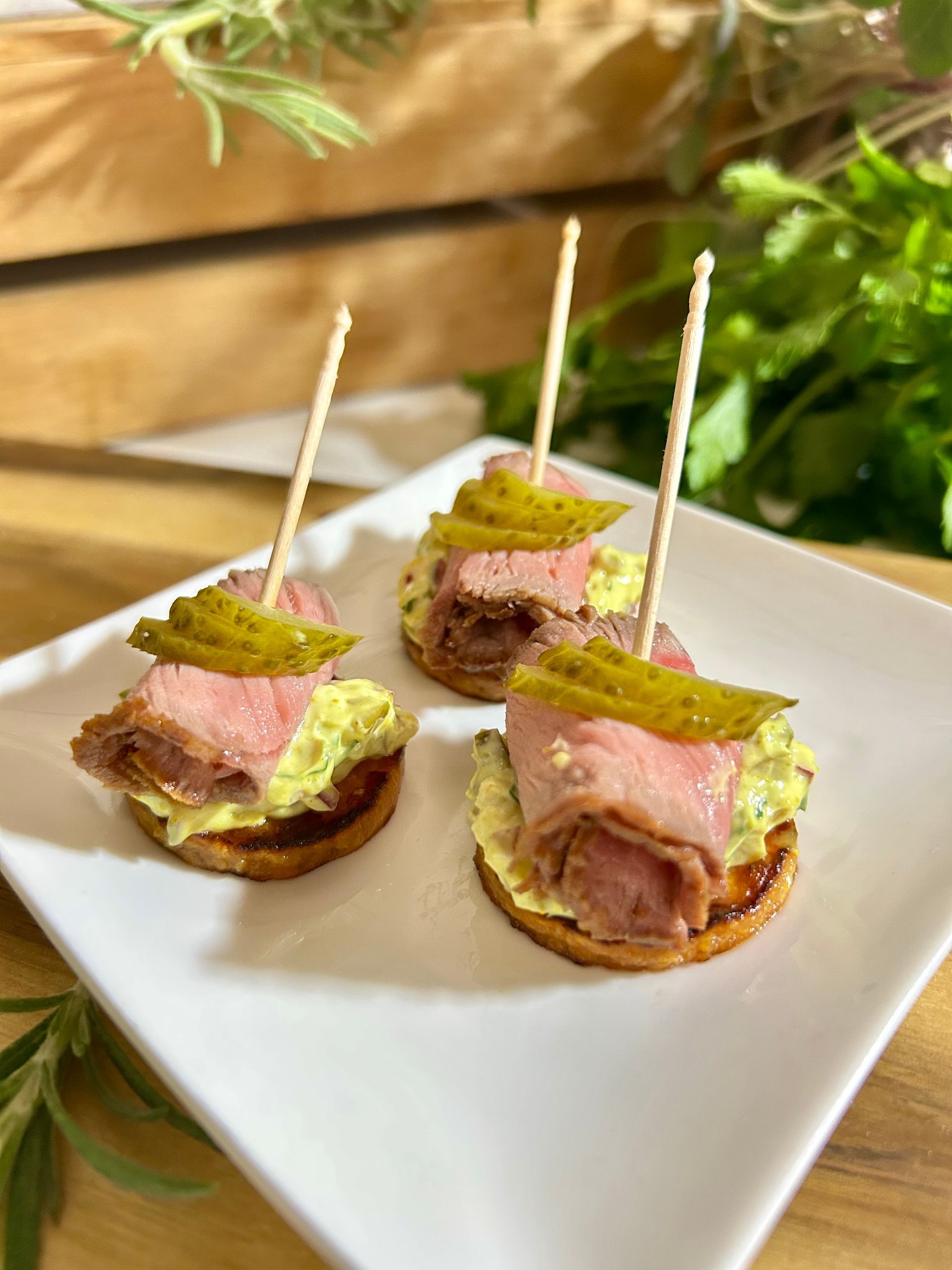 Drei Vorspeisen auf einem weißen quadratischen Teller, jede mit einer Scheibe Roastbeef, einem Klecks Aufstrich und einer Gurkenscheibe auf einem Cracker.