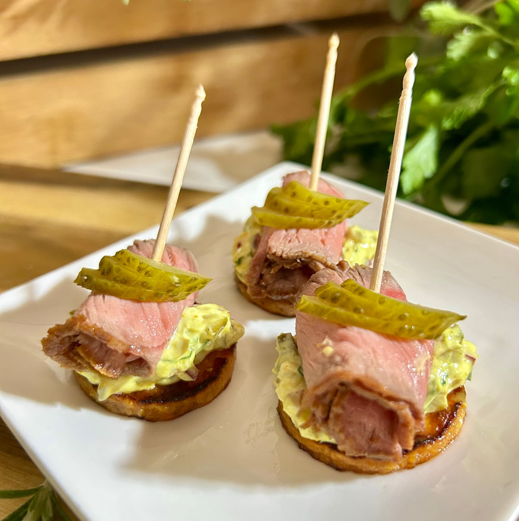 Drei kleine Vorspeisen auf einem quadratischen weißen Teller, jede mit Roastbeef, grüner Soße und einer Gurkenscheibe auf einem Cracker.