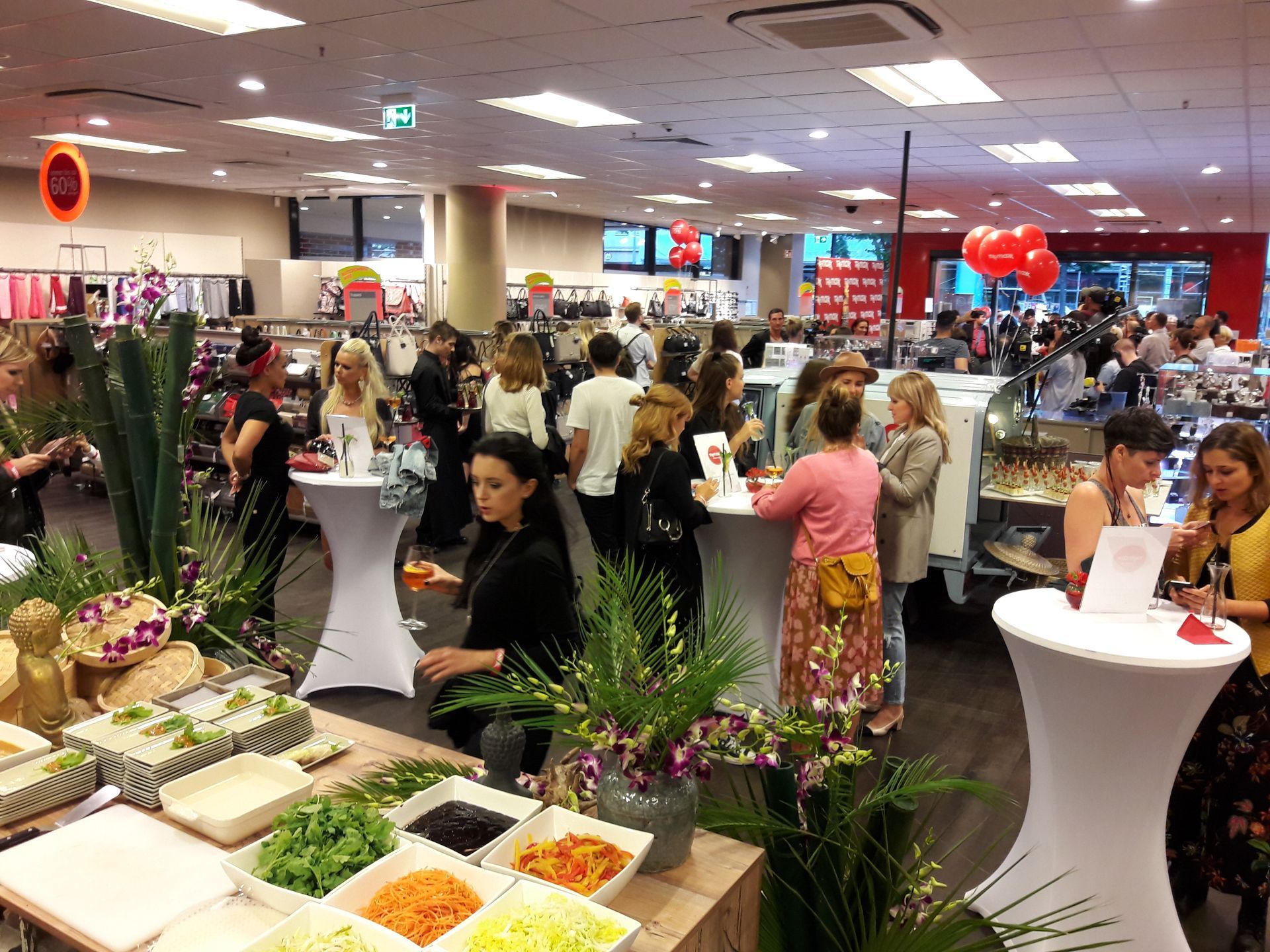 In einem hell erleuchteten, modernen Einzelhandelsgeschäft mit weißen Stehtischen und Blumenarrangements unterhalten sich die Menschen bei einem Buffet.