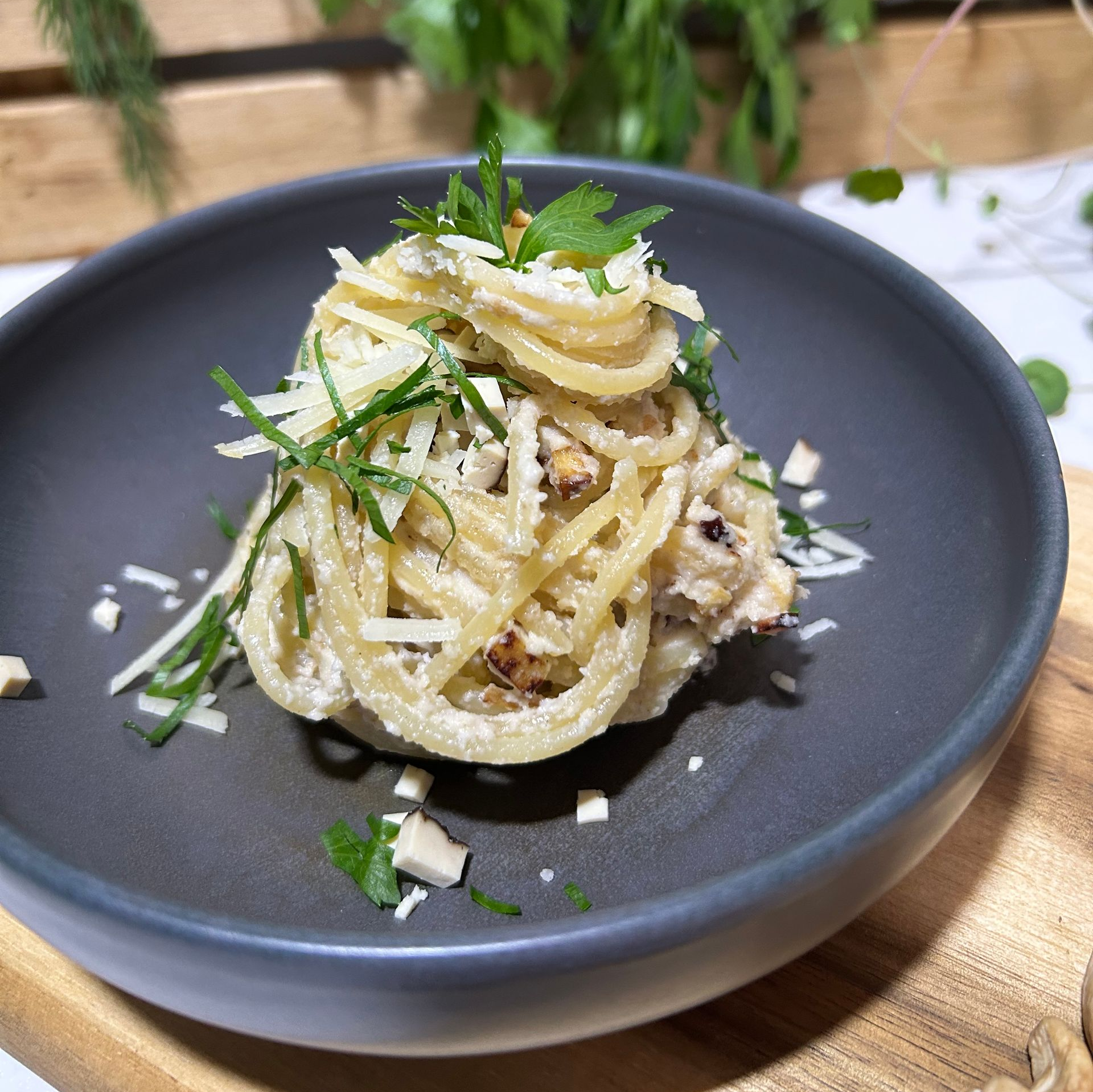 Eine Portion Spaghetti mit geriebenem Käse und gehackten Kräutern steht in einer dunkelgrauen Schüssel auf einer Holzoberfläche.