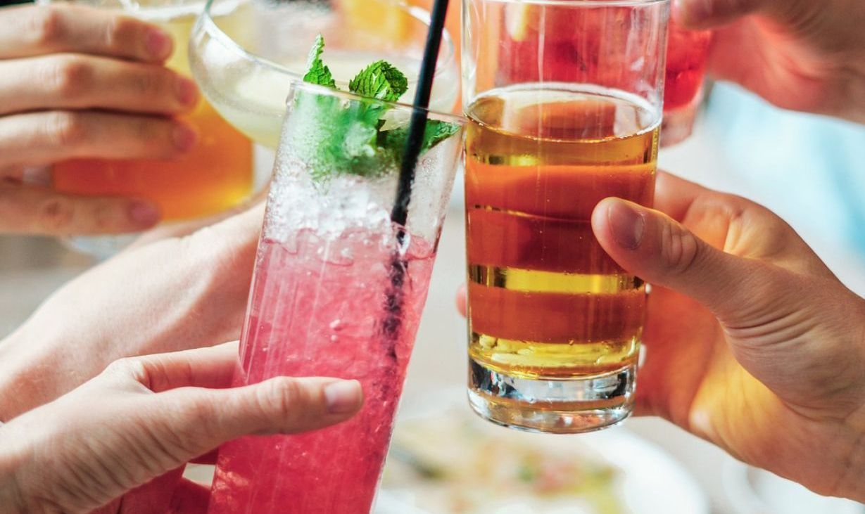 Menschen stoßen mit Cocktails an; rosafarbene, gelbe und bernsteinfarbene Flüssigkeiten in hochgehaltenen Gläsern.