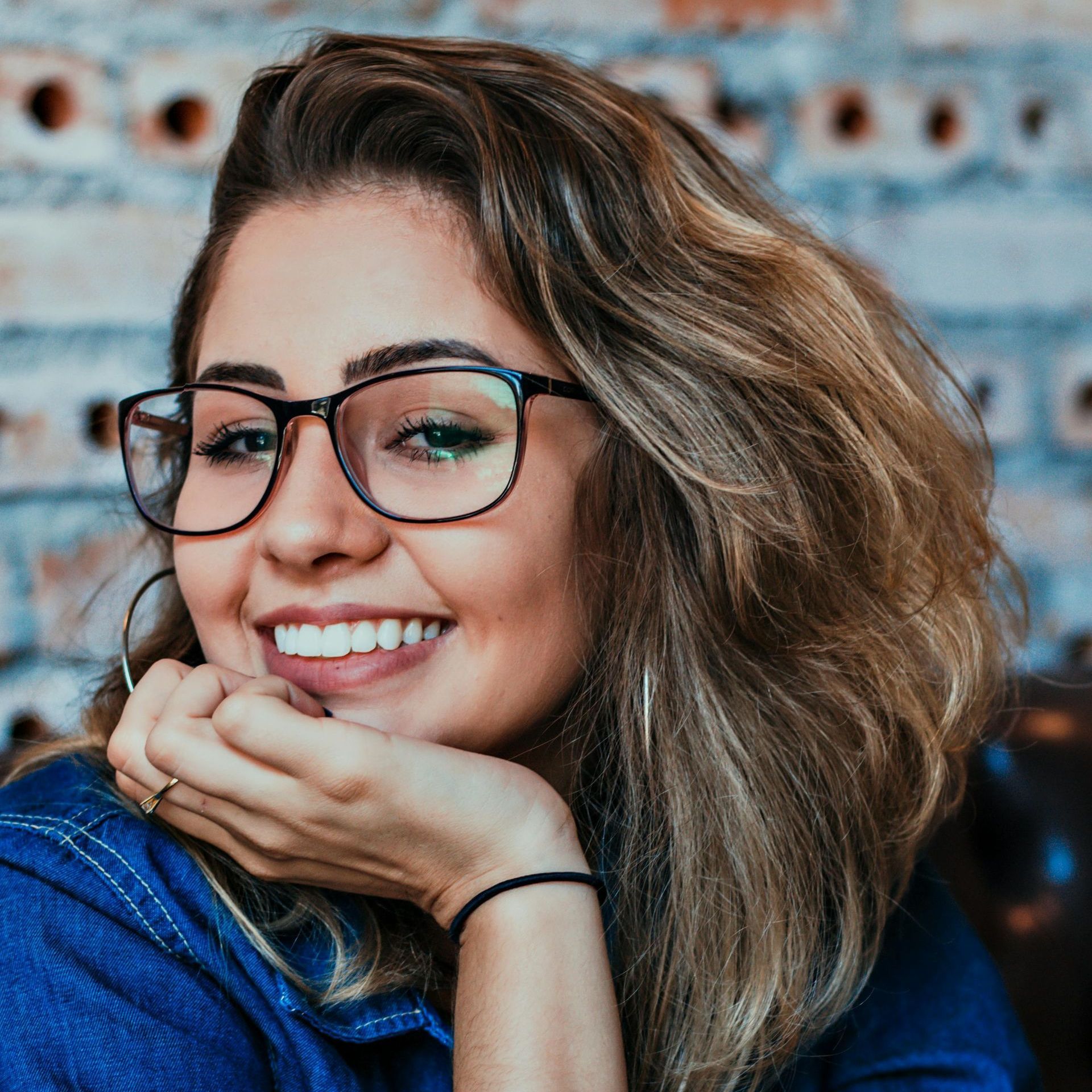 A woman wearing glasses is smiling with her hand on her chin.