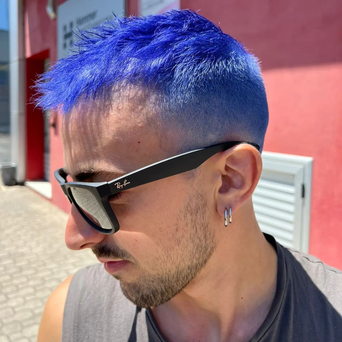 Uomo con i capelli blu e occhiali da sole, che guarda in basso. All'aperto, vicino a un muro rosa.