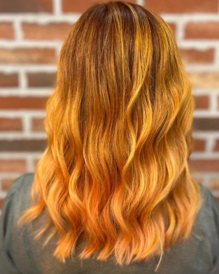 Donna con capelli lunghi e ondulati. I capelli hanno un effetto sfumato dal castano all'arancione brillante, che risalta su un muro di mattoni.
