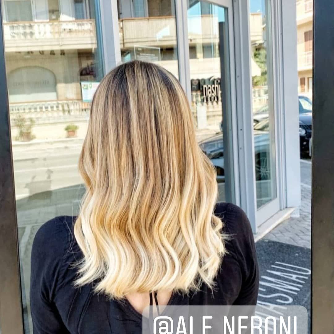 Donna con capelli ondulati e mèches bionde, vista di spalle, di fronte a un edificio con una grande finestra.