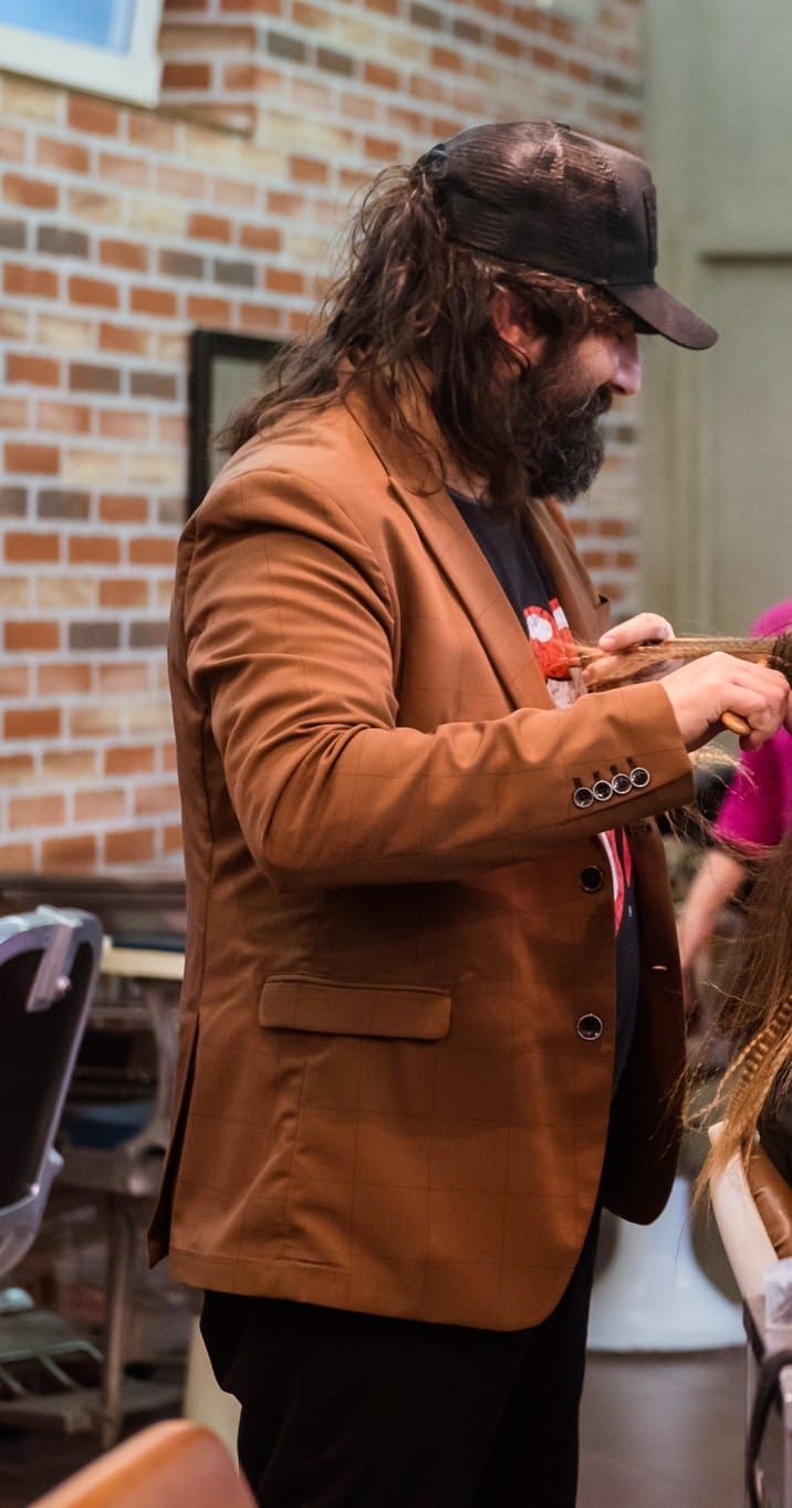 Uomo con giacca marrone, cappello e barba che tiene in mano una tromba, all'interno di una stanza con un muro di mattoni.