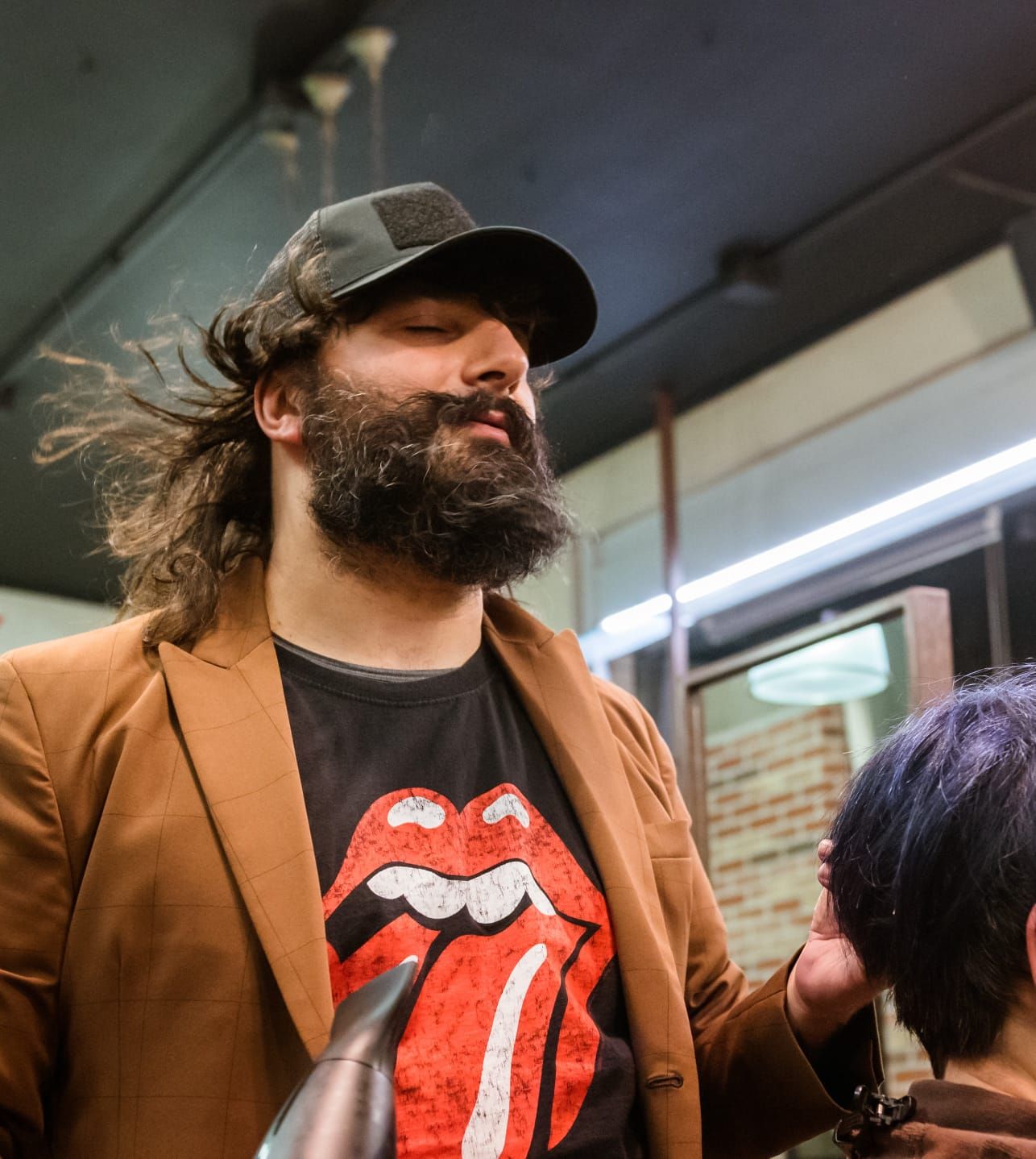 Parrucchiere con barba e cuffia che asciuga i capelli di una cliente, indossando una maglietta dei Rolling Stones e un blazer marrone.
