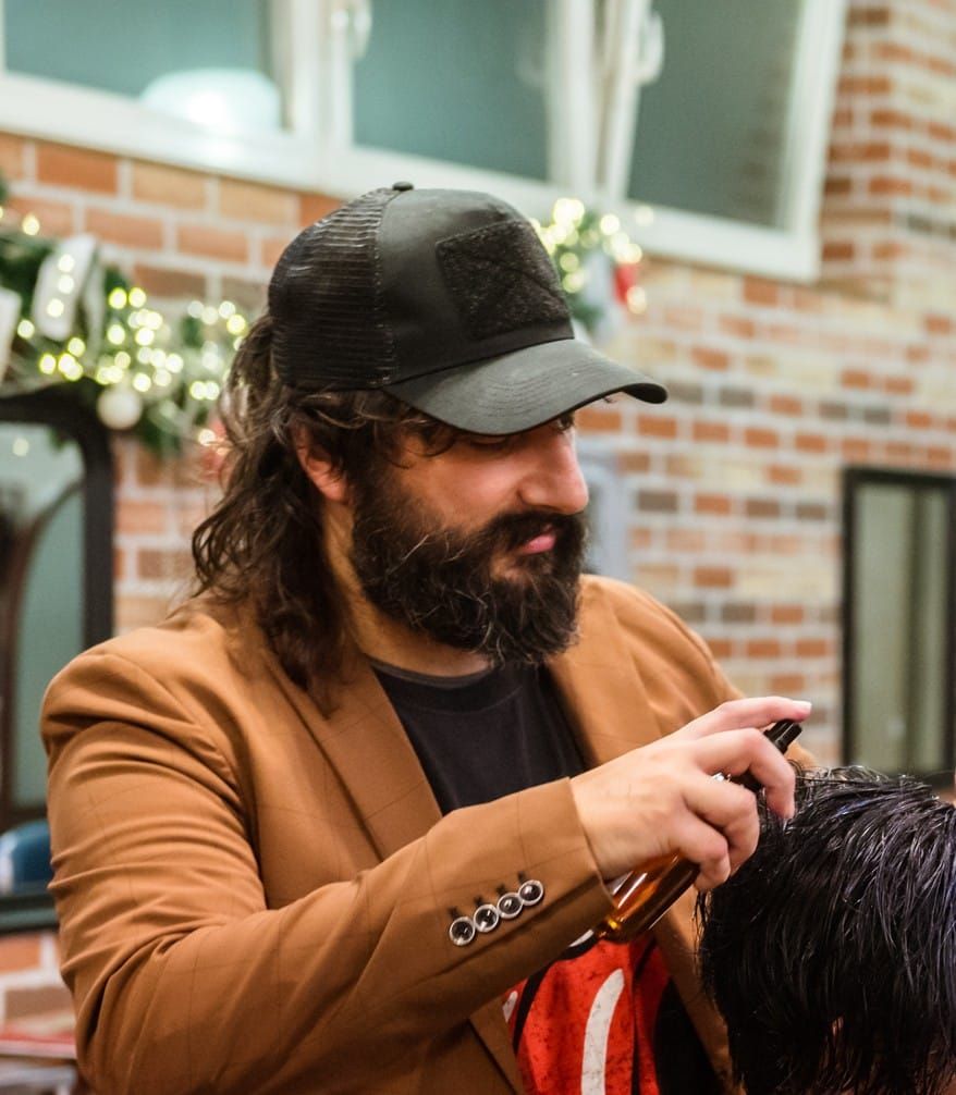 Un uomo con una giacca marrone si lacca i capelli in un salone di bellezza. Indossa un berretto nero e ha la barba.