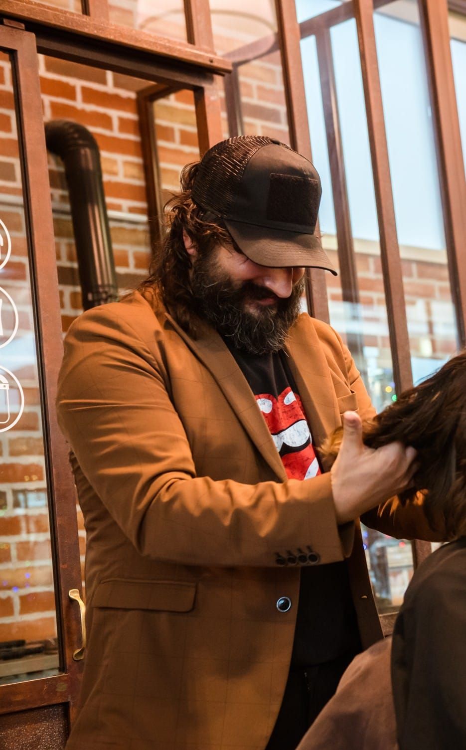 Un uomo con giacca marrone e berretto taglia i capelli in un salone di bellezza; sullo sfondo un muro di mattoni.