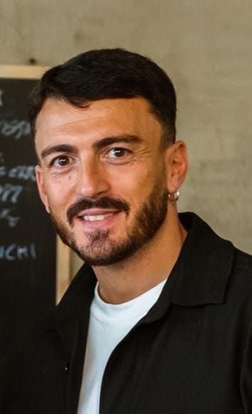 Uomo con barba e capelli scuri, sorridente. Indossa una giacca nera e una camicia bianca, con un orecchino a cerchio.