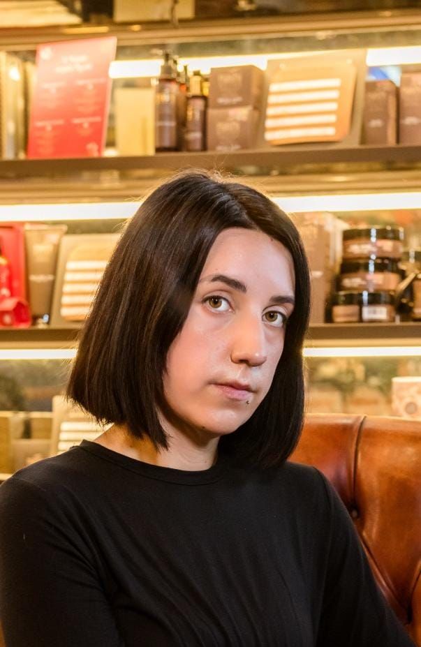 Una donna con i capelli scuri e una camicia nera è seduta su una sedia e guarda avanti in un negozio.