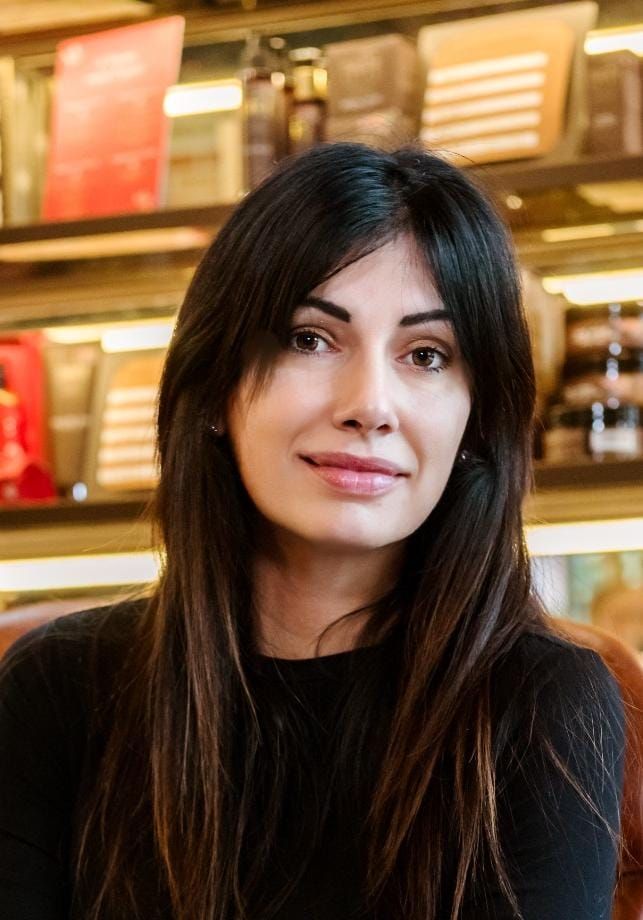 Donna con lunghi capelli scuri e frangia, che indossa un top nero, sorridente, in casa.