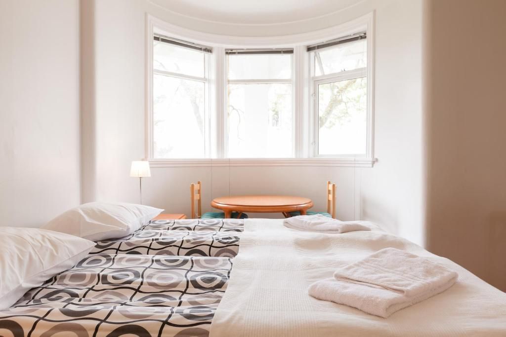 A sunlit bedroom features a bed with patterned linens, a wooden desk under a bay window, and white walls.