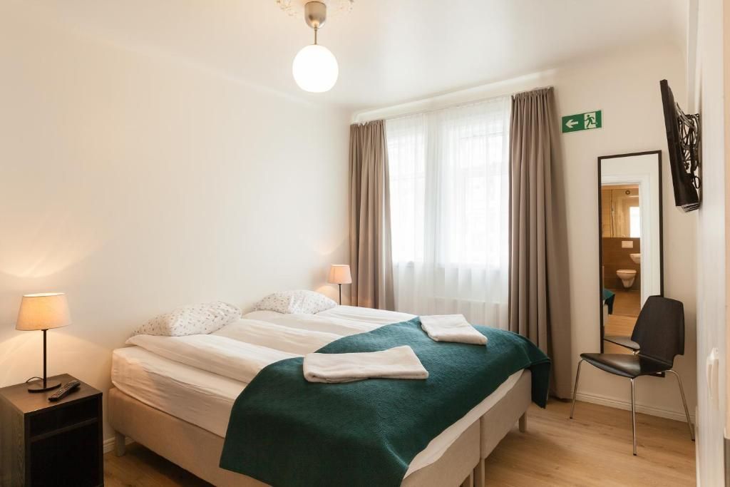 A hotel bedroom with a double bed, bedside lamp, floor mirror, chair, and beige curtains covering a window.