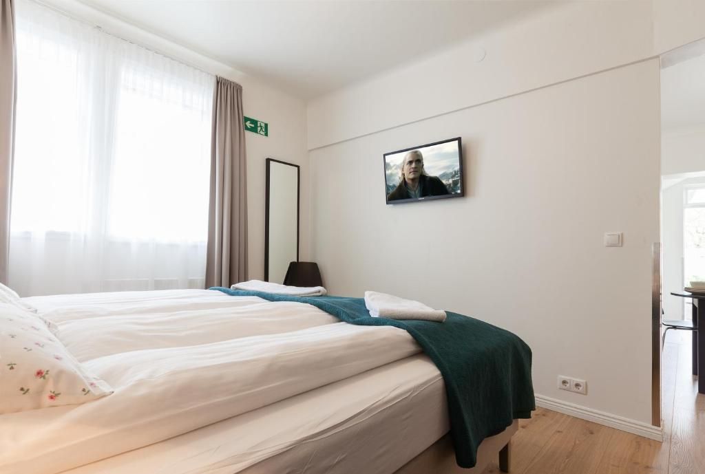 A neat, brightly lit hotel room featuring a white bed with a dark green throw, a wall-mounted TV, and a tall floor mirror.