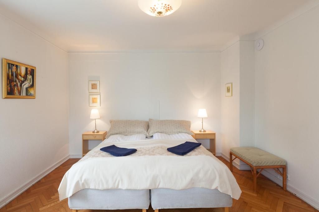 A tidy bedroom with a made bed, two small nightstands with lamps, a bench, and a framed painting on a white wall.