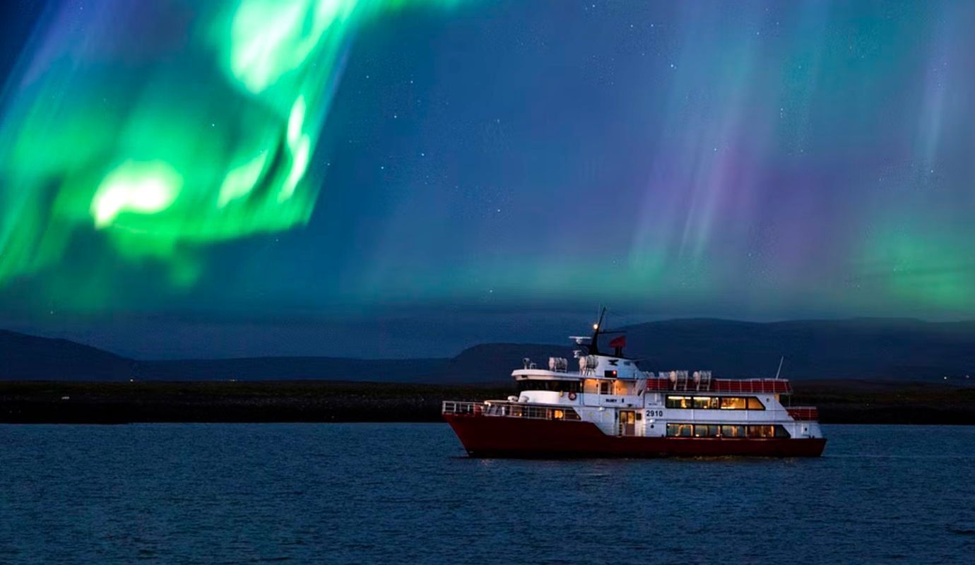 A red and white boat cruises on dark water beneath a vibrant green and purple aurora borealis in a night sky.