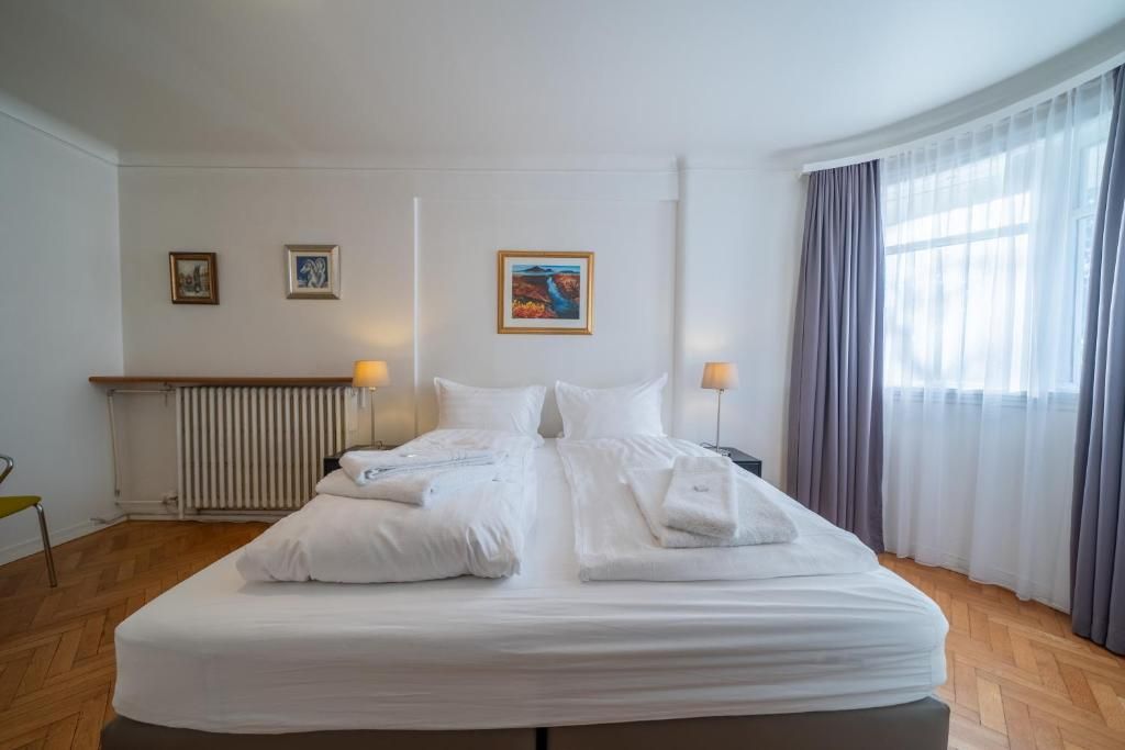 A hotel room featuring a large bed with white linens, wooden herringbone floors, neutral walls, and grey curtains.