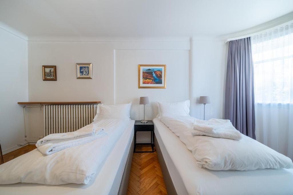 Two single beds with white bedding in a bedroom with wooden floors, neutral walls, and sheer curtains.