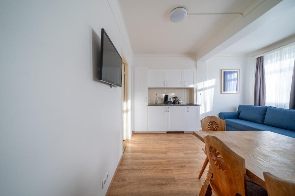 A cozy apartment featuring light wood floors, a blue sofa, a wooden dining table,  a small kitchenette with white cabinets.