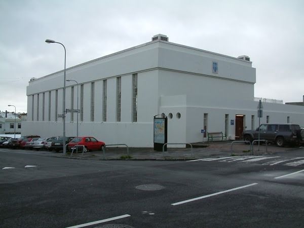 A white, rectangular, modern building with tall, narrow windows and a parking lot, viewed from a street intersection.