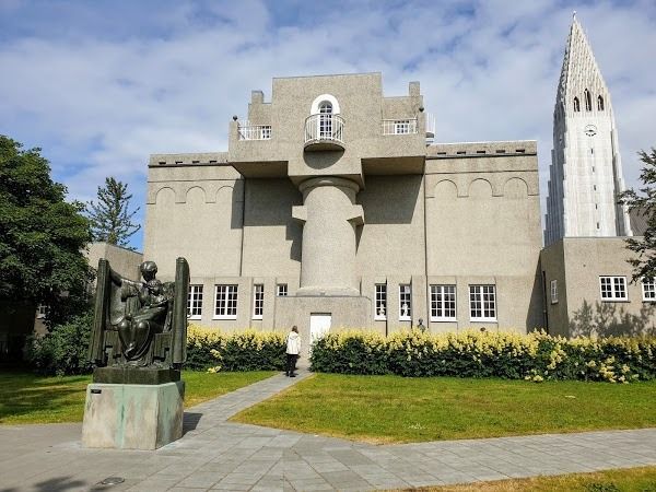 A stone building with a central balcony and pillar, beside a tall, slender tower, with a bronze statue on the lawn.