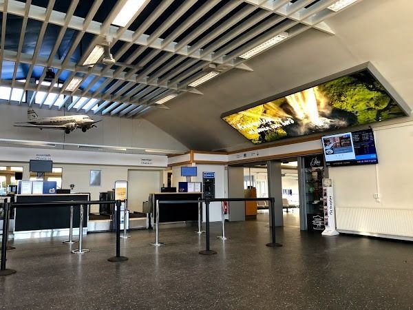 An airport terminal check-in area with black stanchions, counters, an overhead model airplane, and a digital display screen.