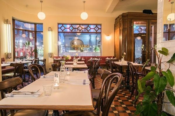 Empty tables and wooden chairs in a bright restaurant with pendant lights and window views of a street at dusk.